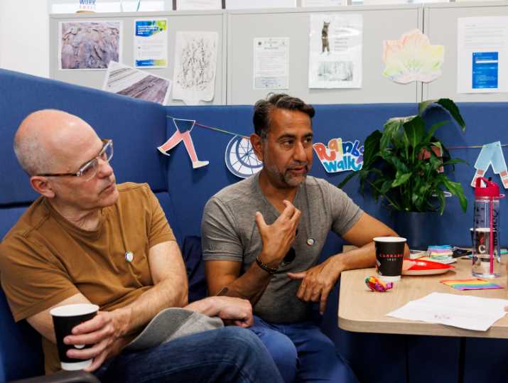 Bill and Irfan, in an orange and grey tshirt and jeans respectively, talk as they sit on a blue sofa, taking part in the Rainbow Walk and Conversation Café.