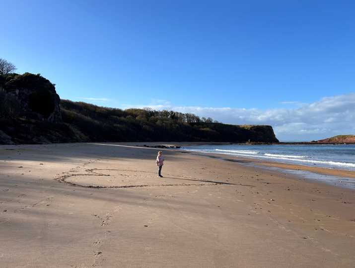 A young girl stands in the middle of a giant heart etched into the sand, with a long beach curving away into the distance below cliffs